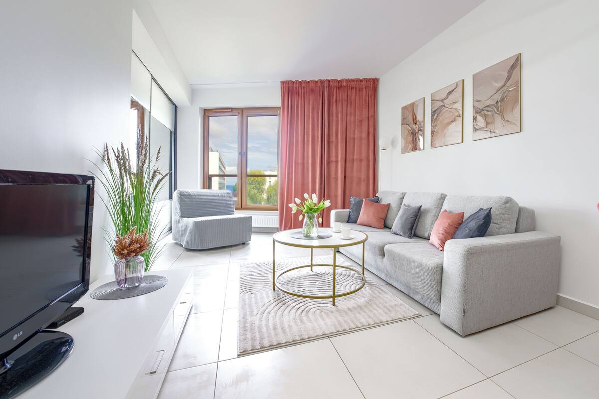 Ein modernes Wohnzimmer mit einem grauen Sofa, einem Couchtisch, einem Fernseher und einem großen Fenster mit Vorhängen.