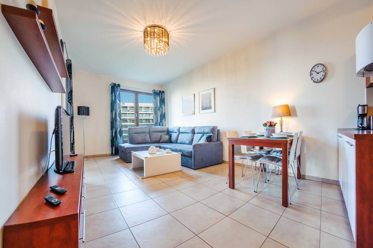 Ein modernes Wohnzimmer mit Sofa, Couchtisch und Essbereich. Ein Fernseher steht auf einem Schrank, eine Lampe und eine Uhr sind an der Wand.