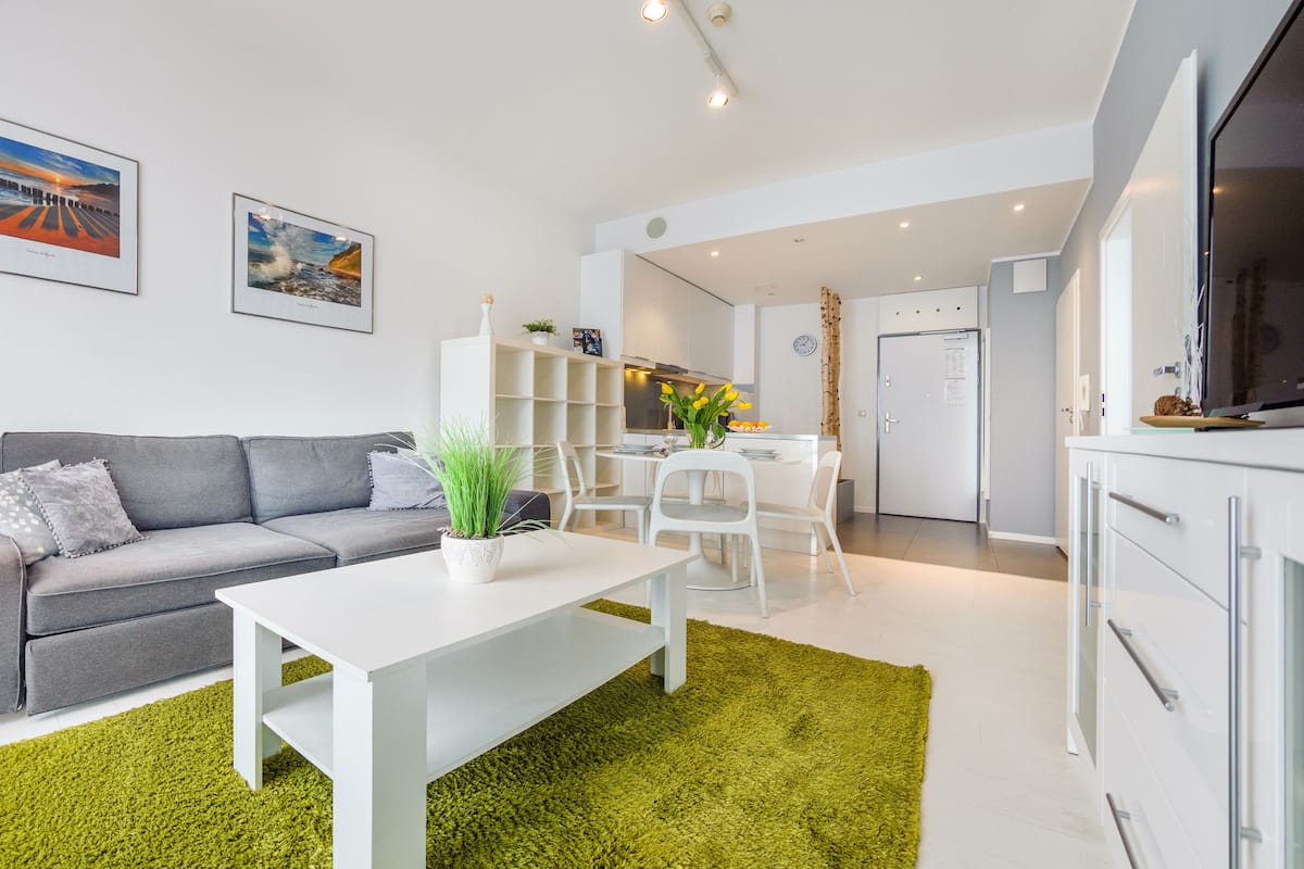 Ein modernes Wohnzimmer mit grauem Sofa, weißem Couchtisch und grünem Teppich. An der Wand hängen zwei gerahmte Bilder, ein Küchenbereich mit weißen Schränken und ein Esstisch mit Stühlen.