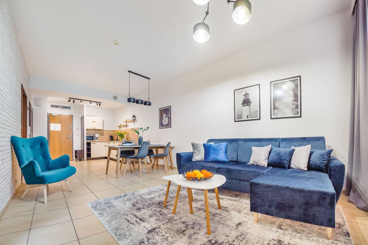 Ein modernes Wohnzimmer mit einem blauen Sofa, einem blauen Sessel, einem Esstisch und einer Küche im Hintergrund.