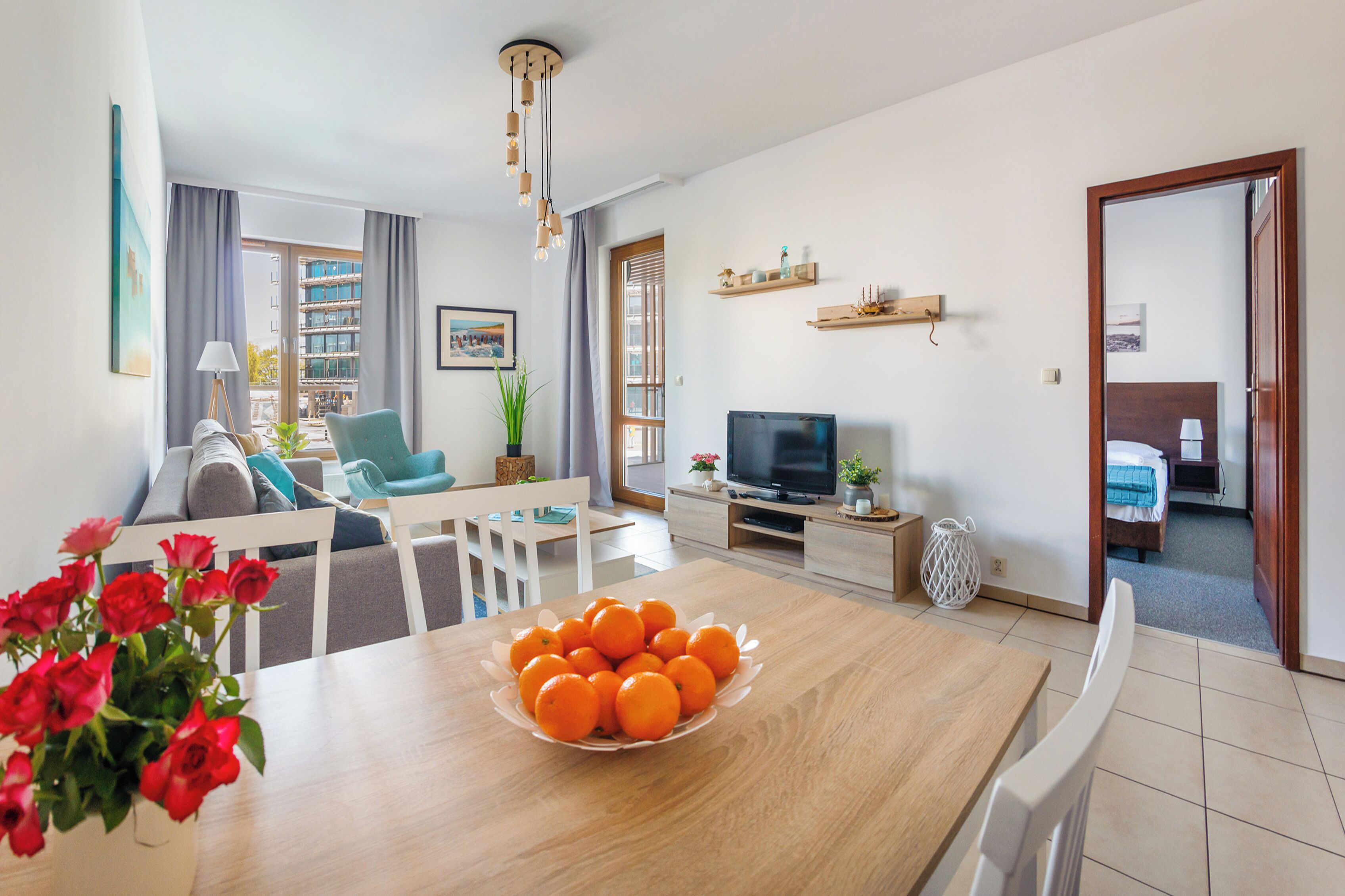 Una sala de estar moderna con una mesa de comedor de madera, un televisor y un sofá. Se ve un balcón a través de la ventana.