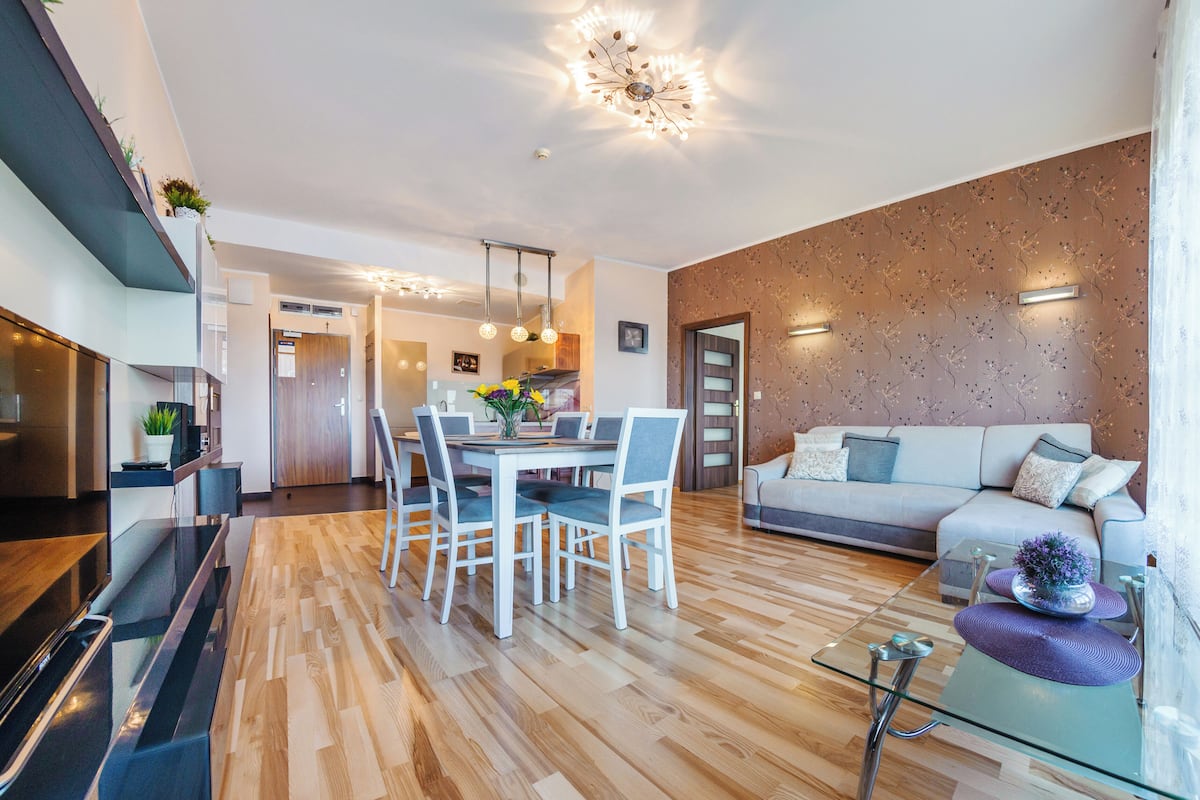 Ein modernes Wohnzimmer mit Essbereich, Holzboden, einem Glastisch und einer Couch mit dekorativen Kissen.