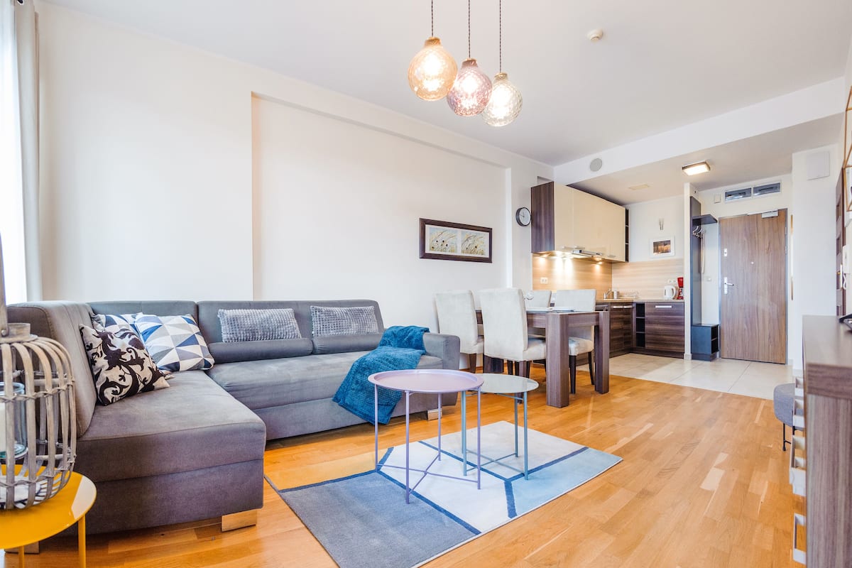 Ein modernes Wohnzimmer mit einem grauen Sofa, einem kleinen runden Tisch und einem blauen Teppich. Im Hintergrund ist eine Küchenzeile mit Holzschränken und einem Esstisch zu sehen.