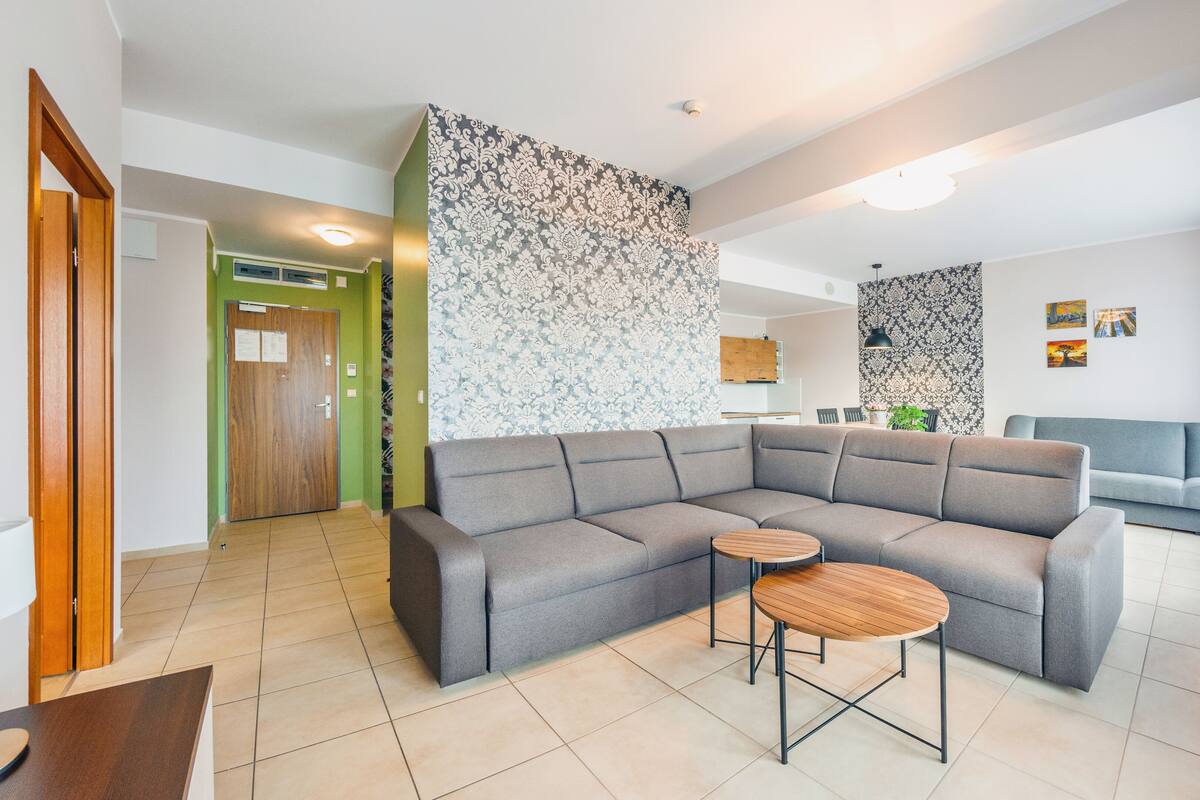 Ein modernes Wohnzimmer mit einem grauen Ecksofa, einem runden Couchtisch aus Holz und Wandpapier mit Muster.