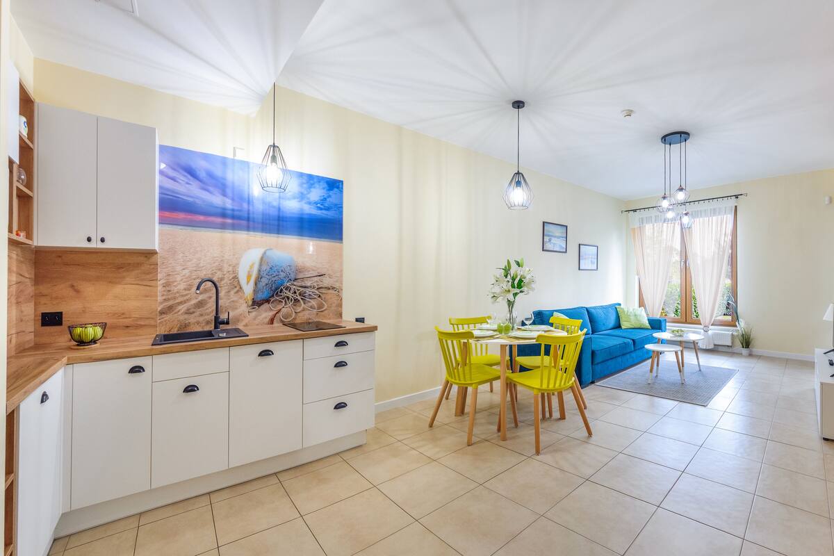 Eine moderne Küche mit weißen Schränken, ein Essbereich mit gelben Stühlen, ein blaues Sofa und ein großes Fenster mit Vorhängen.