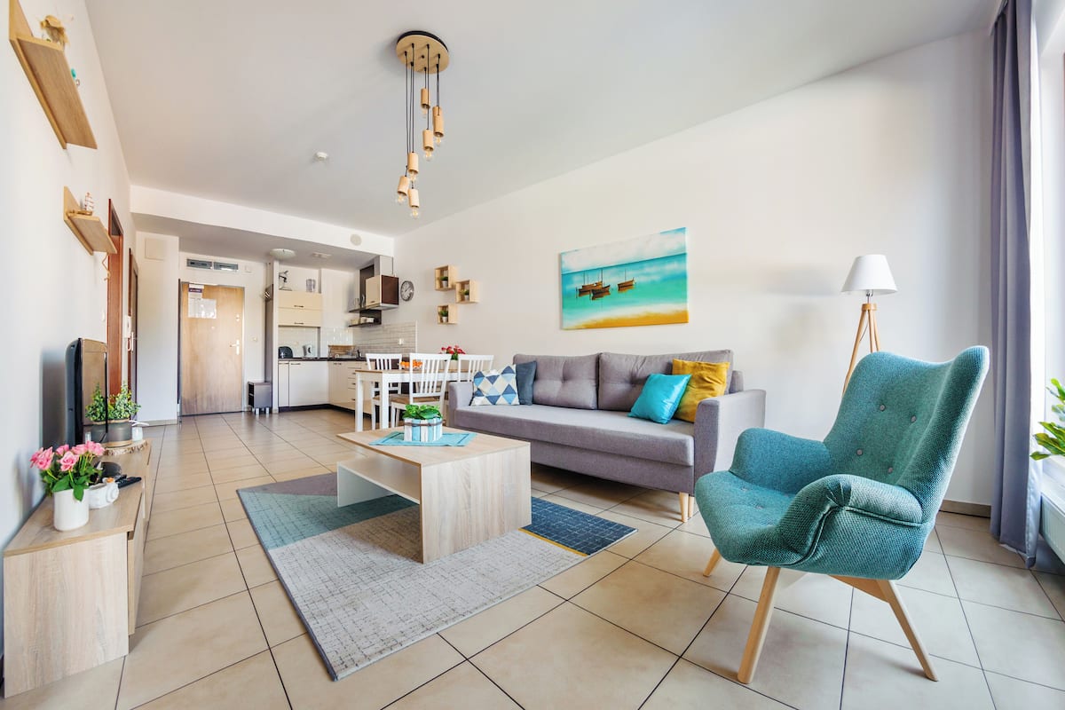 Ein modernes Wohnzimmer mit einer Couch, einem Couchtisch und einem Türkis-farbenen Sessel. Im Hintergrund ist eine Küche zu sehen und an der Wand hängt ein Gemälde.