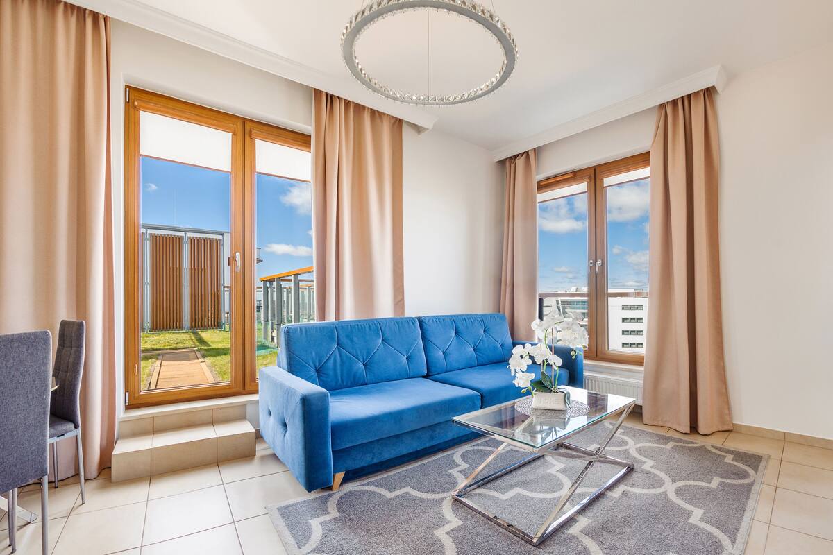 Ein modernes Wohnzimmer mit einem blauen Sofa, einem Glastisch und Blick auf einen Balkon mit Holzzaun.
