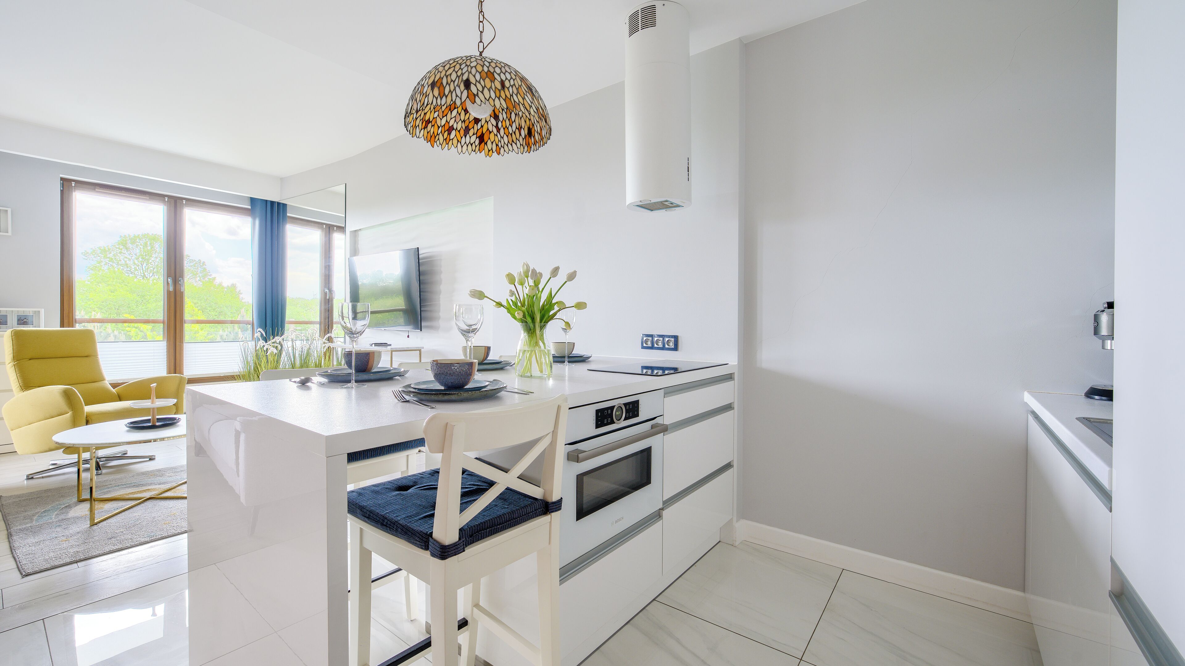 Una cocina moderna con armarios blancos, una mesa de comedor con sillas, un ventanal con vistas a zonas verdes y una lámpara colgante original.