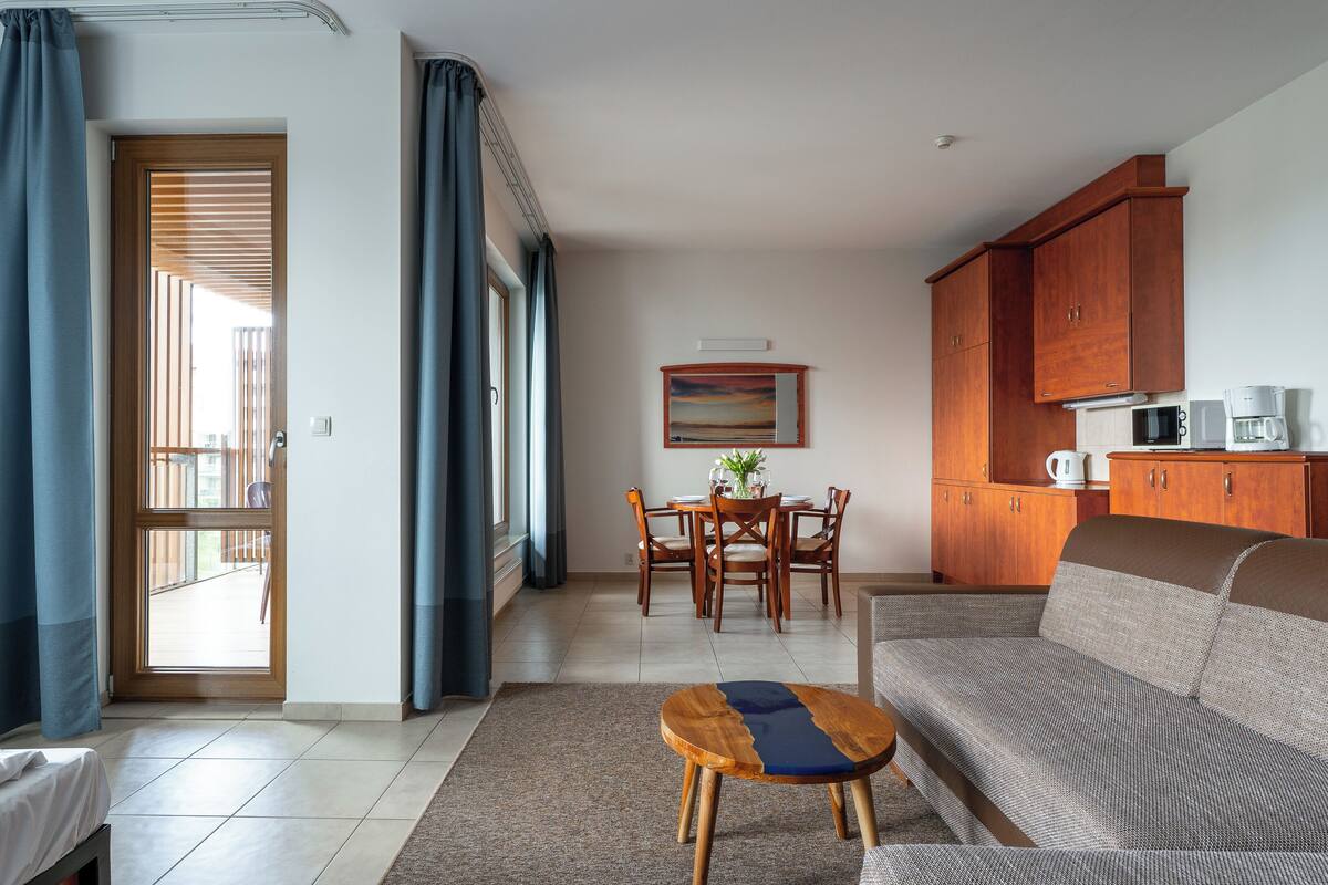 Ein modernes Wohnzimmer mit einer Couch, einem Couchtisch und einem Essbereich mit Tisch und Stühlen. Es gibt eine Küchenzeile mit Schränken und Geräten.