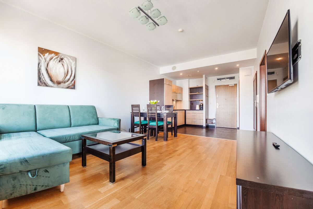 Ein modernes Wohnzimmer mit einem türkisfarbenen Sofa, einem Glastisch und einem Essbereich mit Holzstühlen und einem Tisch.