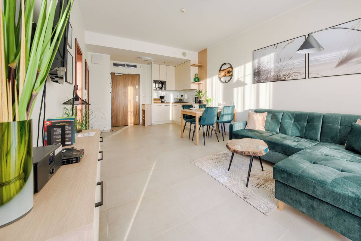 Ein modernes Wohnzimmer mit einem türkisfarbenen Sofa, einem Essbereich mit einem Holztisch und einer Küche im Hintergrund.