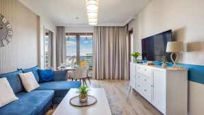 A modern living room with a blue sofa, a dining table with chairs, a kitchen area with a TV, and a view of the city. - Sun & Snow - Olympic Park (Kolobrzeg)