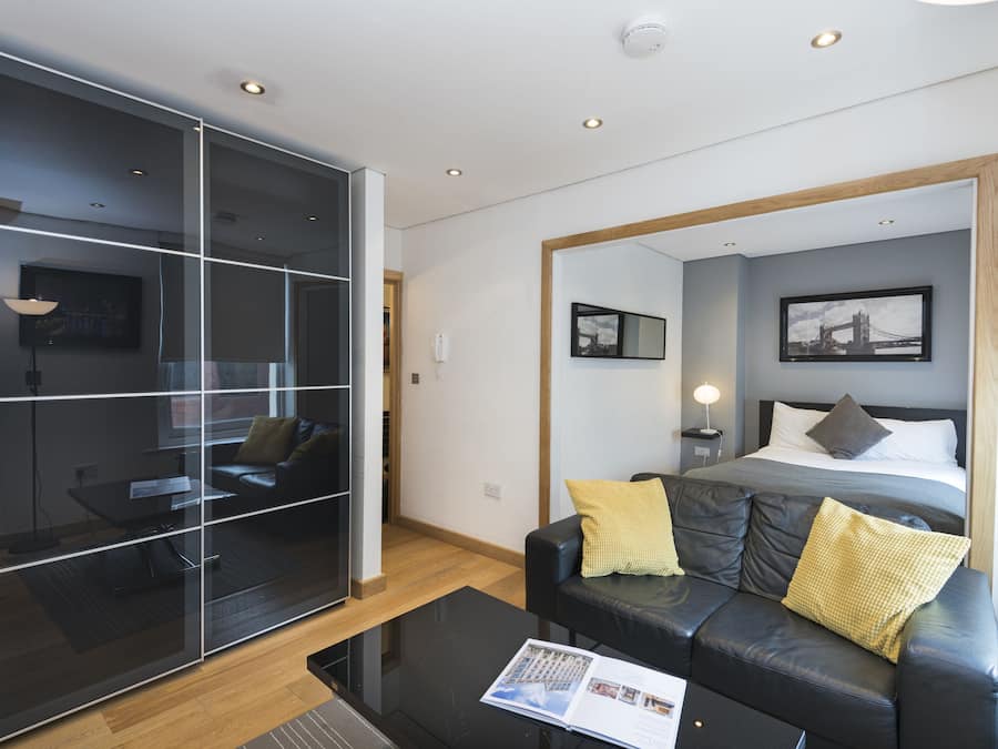 A modern hotel room with a large bed, a leather sofa, a glass partition, and a view of the cityscape.
