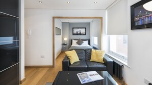 Une chambre d’hôtel moderne avec un canapé en cuir noir, un lit avec du linge de lit blanc, un tableau encadré au mur et une fenêtre donnant sur un panorama urbain.