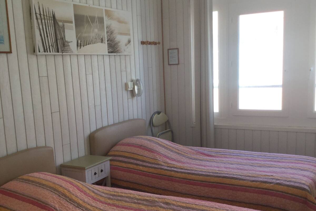Een kamer met twee bedden, een klein nachtkastje, een wandplank met decoratieve objecten en een raam met gordijnen.