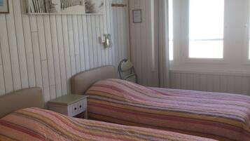 A room with two beds, a small bedside table, a wall-mounted shelf with decorative items, and a window with curtains.