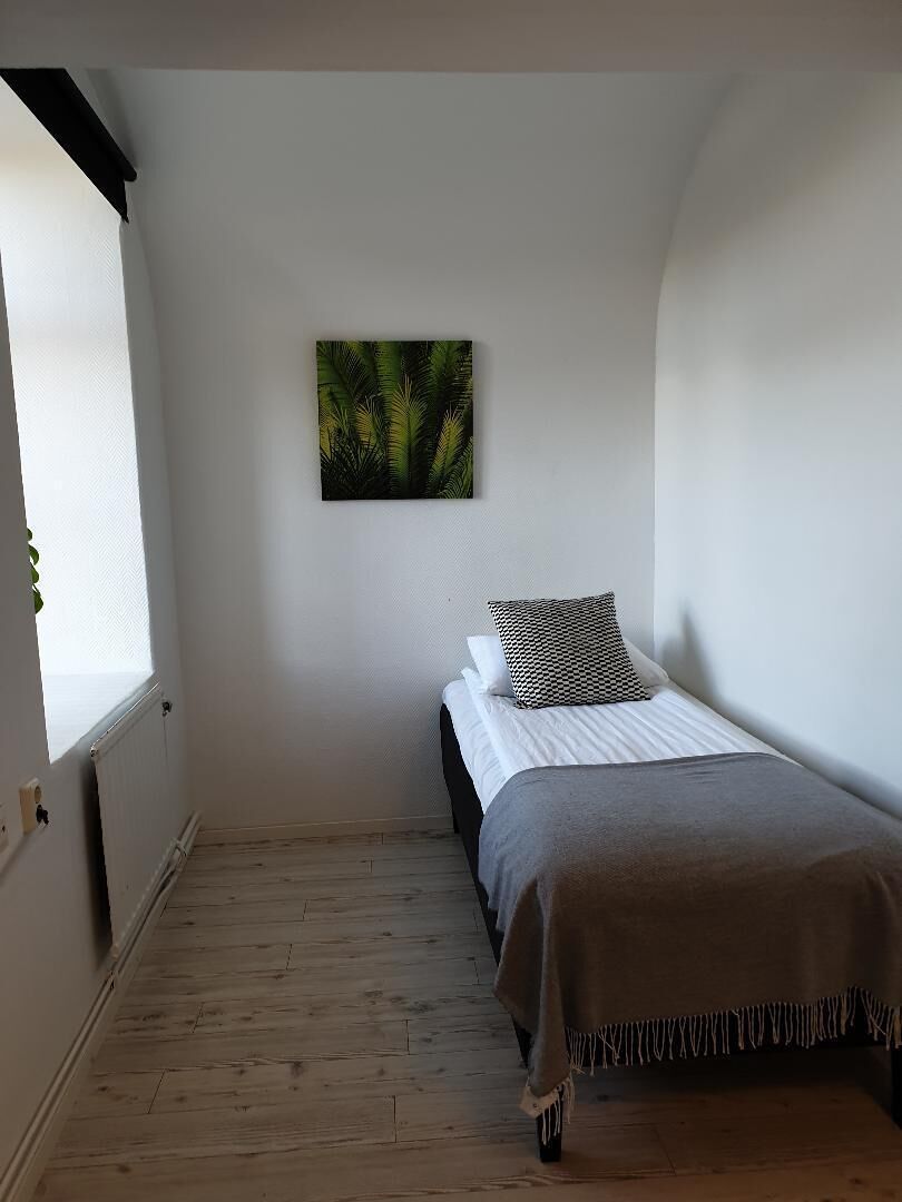 A small, single-bed room with a white wall, a framed picture, and a window on one side.