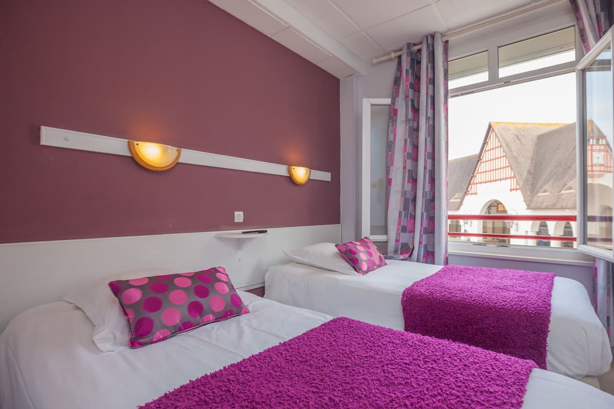Une chambre d’hôtel avec deux lits, chacun agrémenté de linge de lit blanc et de touches de couleur violette. On y trouve des lampes fixées au mur et une vue sur un bâtiment à l’extérieur.