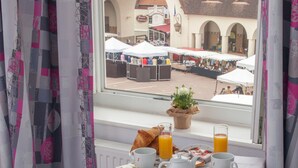 Ein Hotelzimmer mit Frühstückstisch, gedeckt mit Croissants, Kaffee, Tee und Saft. Ein Fenster bietet Blick auf die Straße und ein Gebäude mit Bögen.