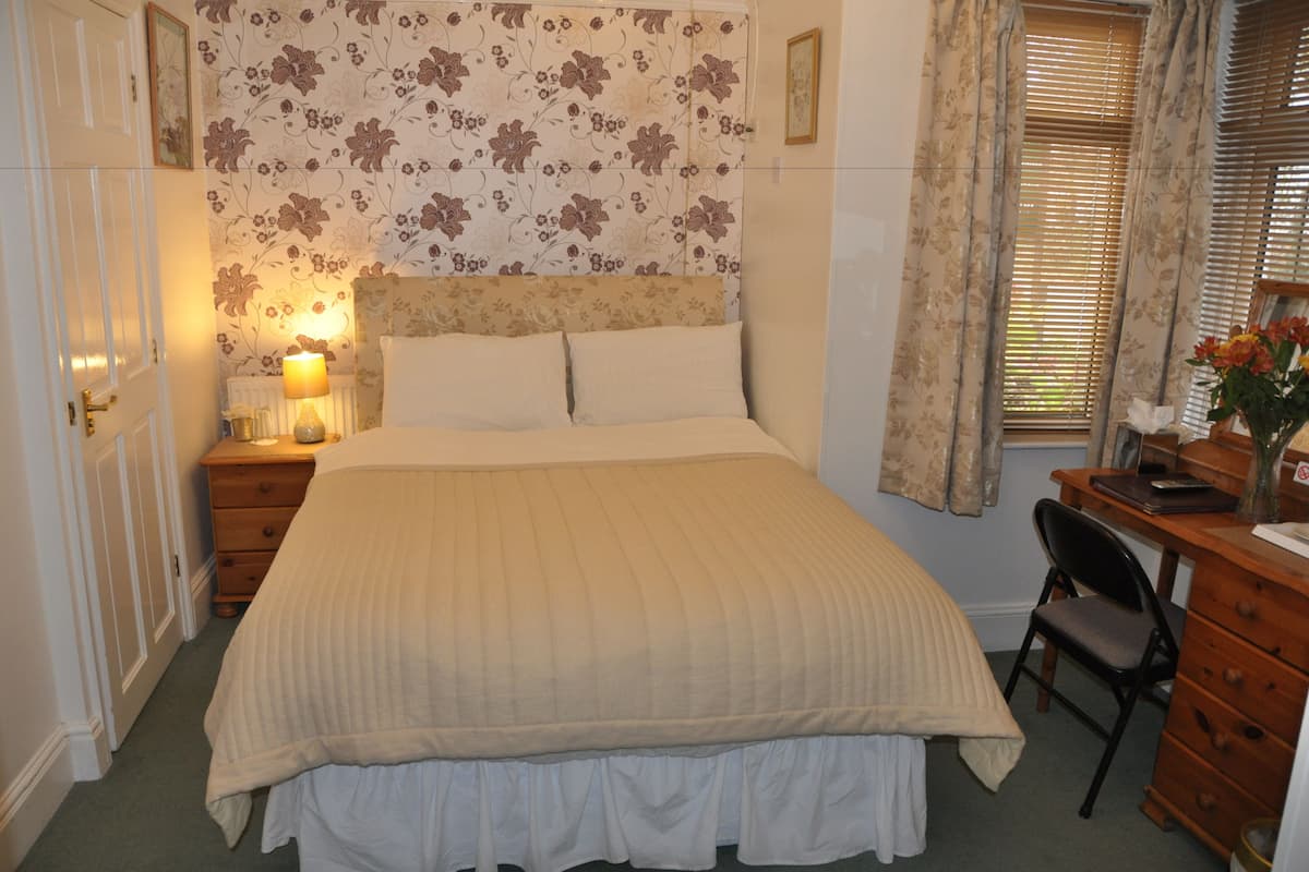A bedroom with a bed, a nightstand with a lamp, a wooden dresser, a desk with a chair, and a window with curtains.
