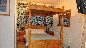 Una cama con dosel de madera, acompañada de una mesita de noche y un aparador a juego, un radiador y papel tapiz con motivos florales.
