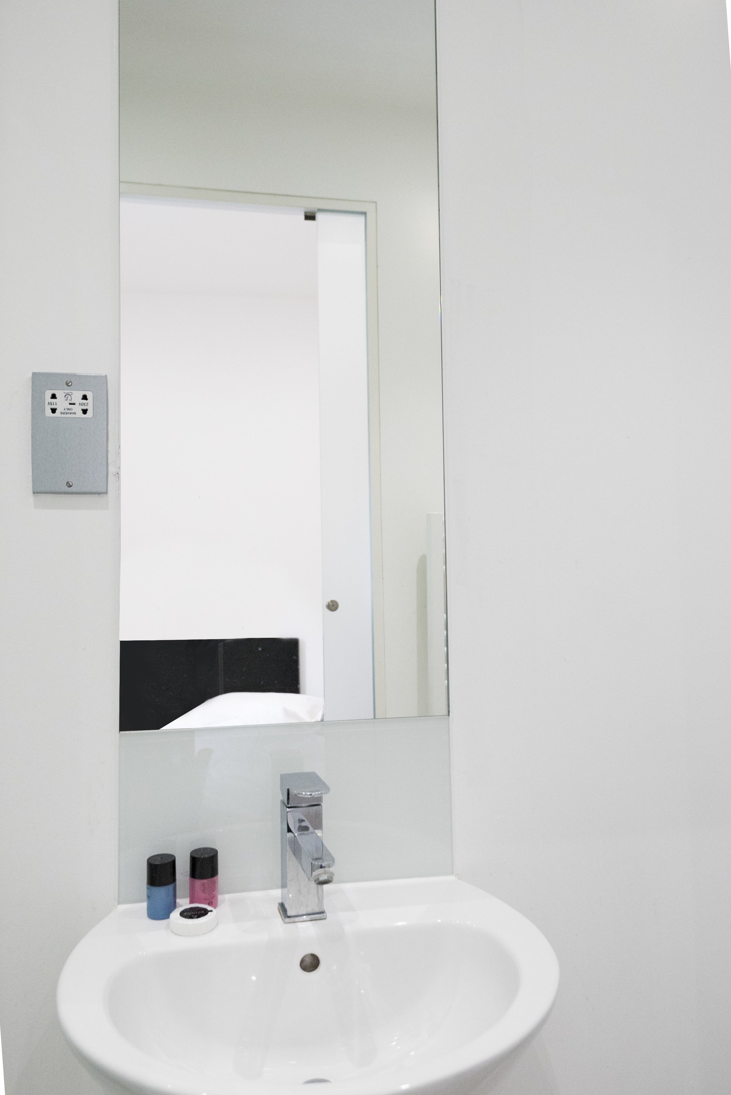 A bathroom with a white sink, a mirror, and a door. There are two bottles on the sink and a light switch on the wall.