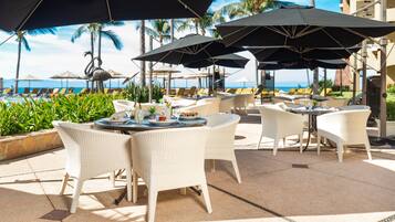 An outdoor dining area with white wicker chairs and tables, large umbrellas, and a view of the ocean.