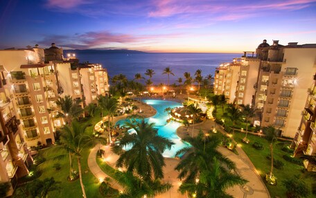 A resort with a swimming pool surrounded by palm trees and a view of the ocean at sunset.