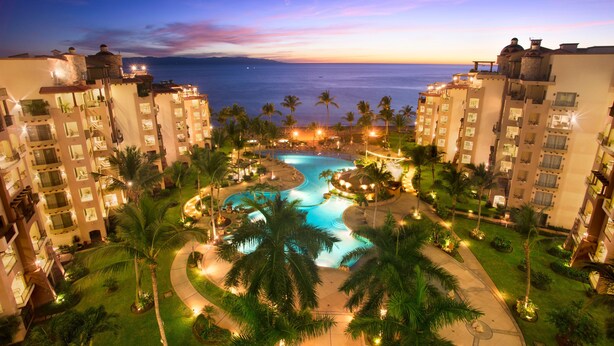 Un resort con piscina rodeada de palmeras y vista al océano al atardecer.