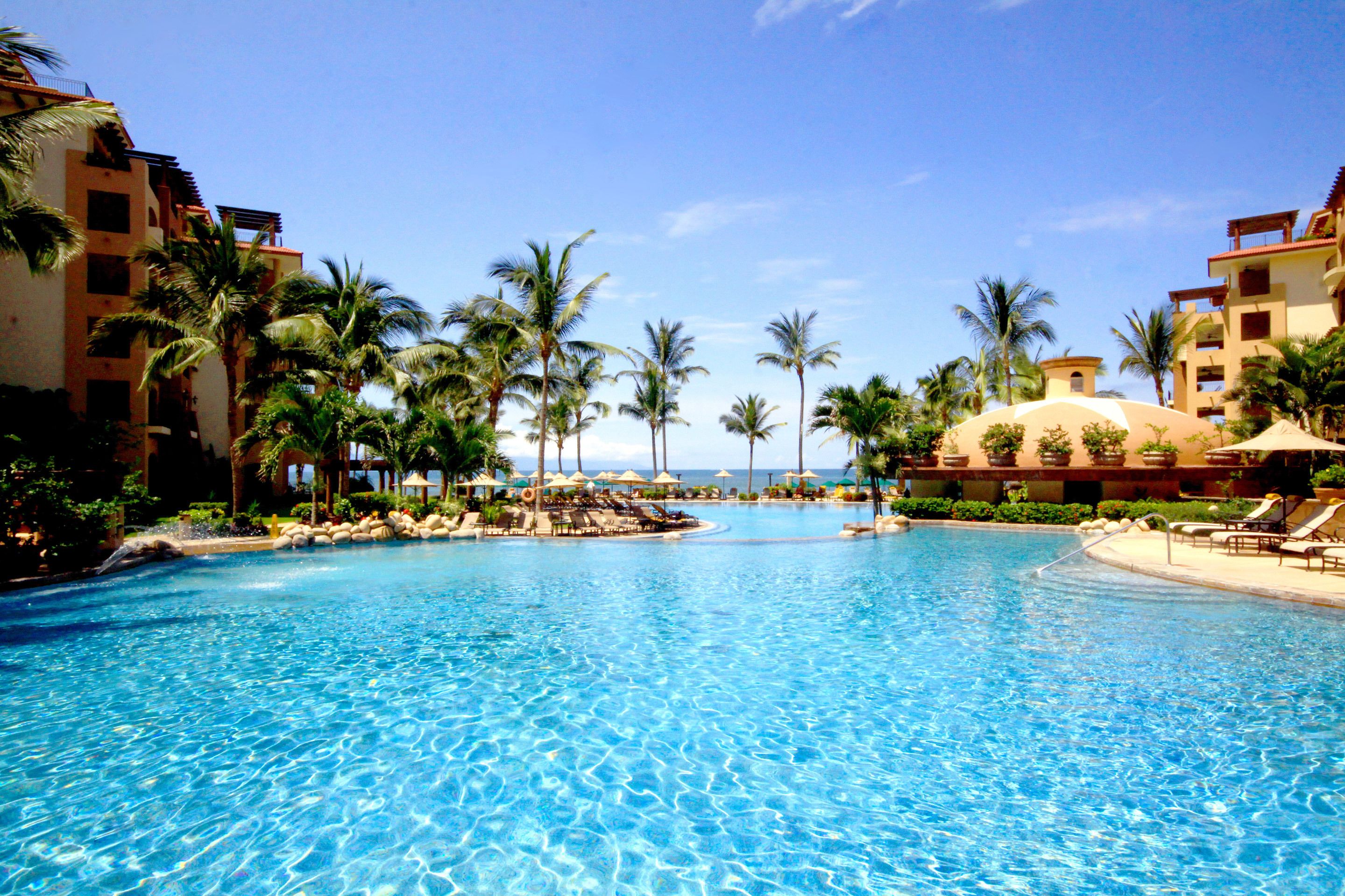 A large swimming pool surrounded by palm trees and resort buildings.