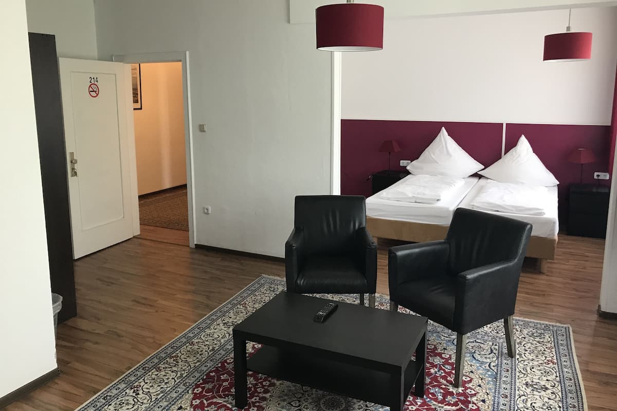 A hotel room with a bed, two black chairs, a small table, a remote control, and a patterned rug.