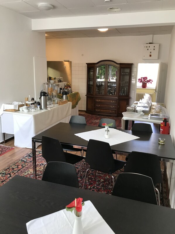 A dining area with a long table, black chairs, and a white tablecloth. There is a cabinet with glass doors and a fire extinguisher on the wall. - Garni Hotel Lore (Hamburg)