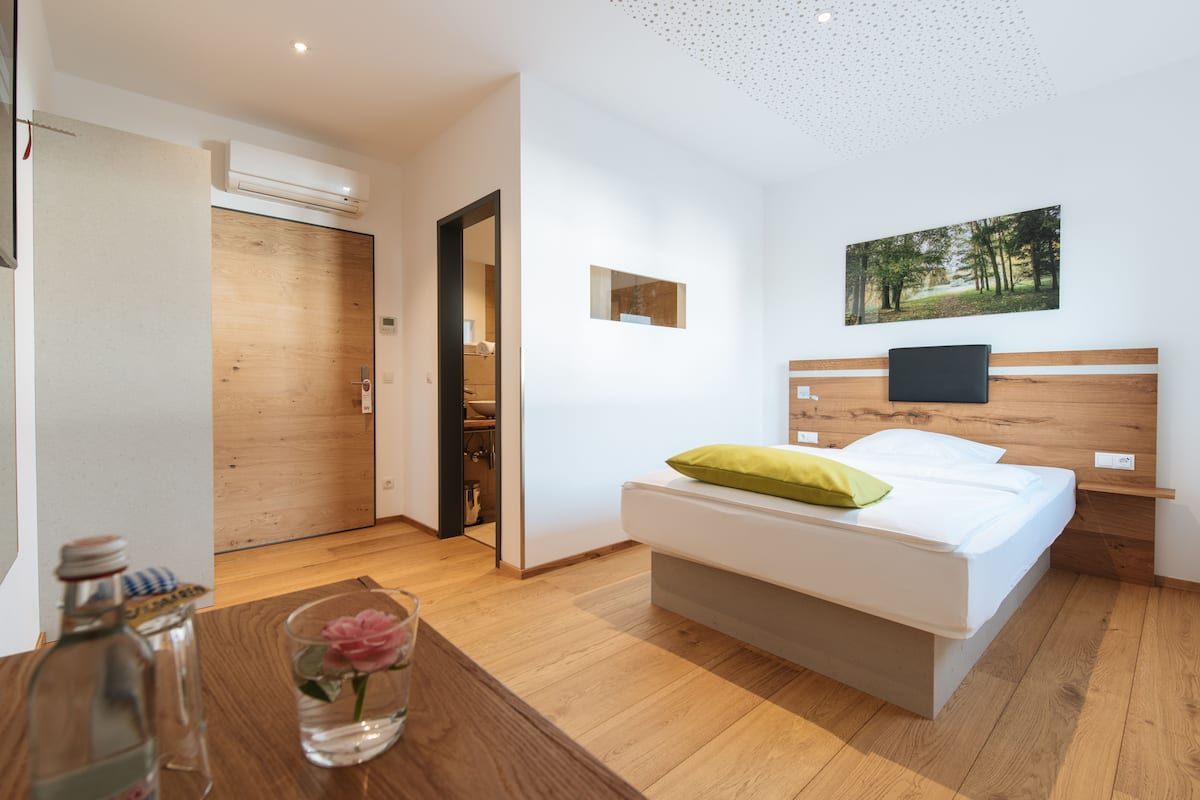 A modern bedroom with a wooden floor, a bed with white bedding and a green pillow, a mounted TV, and a framed picture on the wall.