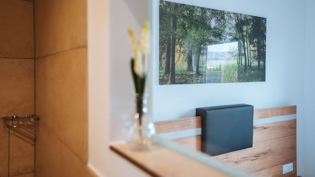 A bathroom with a wooden bench, a framed picture of a forest, and a vase with flowers. - Aparthotel Nah dran (Loiching)