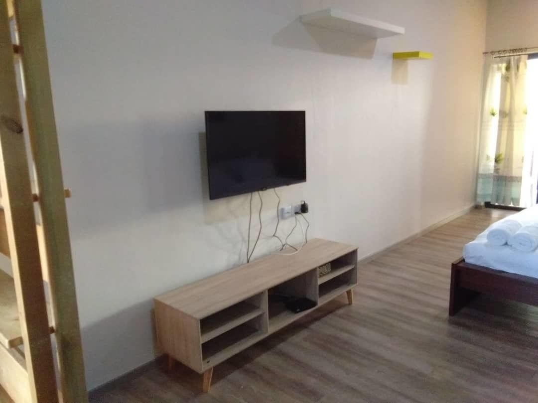 Habitación con una televisión de pantalla plana montada en la pared, un mueble de televisión de madera con estantes, una cama con edredón blanco y una ventana con cortinas.