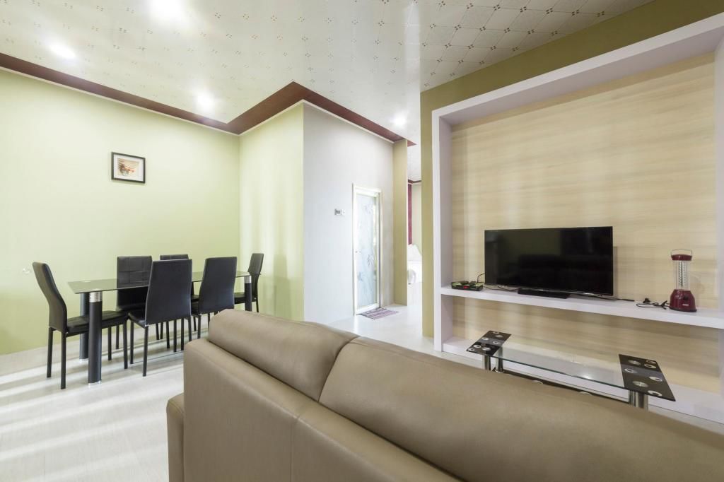 Un salon moderne avec un canapé beige, un coin repas comprenant une table et des chaises, un téléviseur à écran plat posé sur une étagère en bois, et une porte donnant sur une autre pièce.