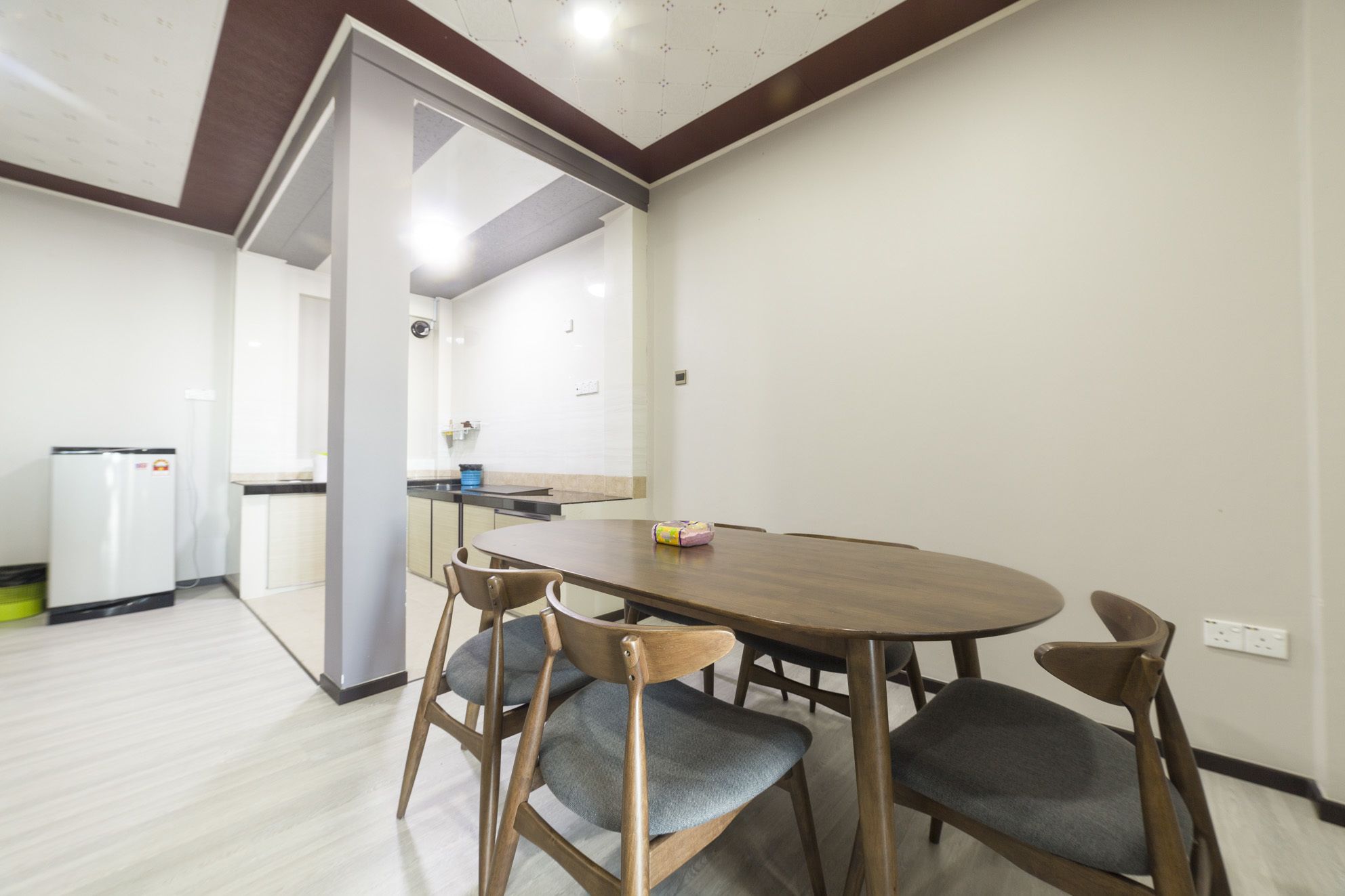 A dining area with a wooden table and four chairs, a refrigerator, and a kitchen area in the background.