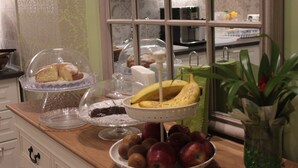 Una encimera de cocina con diversos alimentos, incluyendo un frutero con plátanos y manzanas, un pastel sobre un expositor de vidrio, y un cuenco de cristal con un postre.