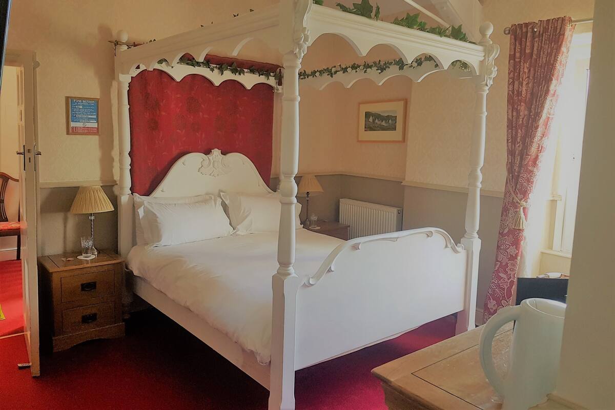 A four-poster bed with white bedding, a nightstand with a lamp, a radiator, and a window with curtains.