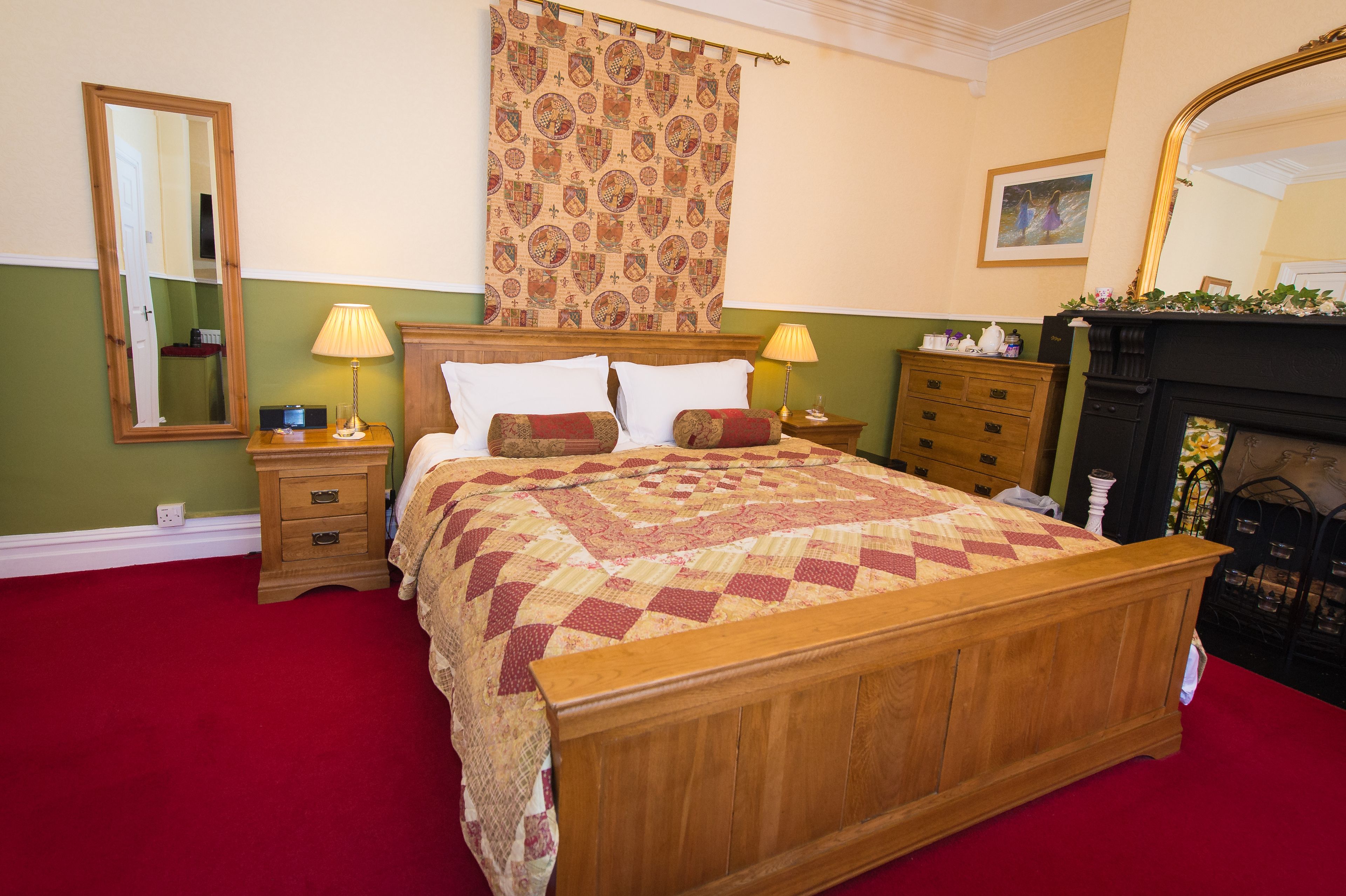 A traditional bedroom with a wooden bed, two bedside tables with lamps, a mirror, a patterned tapestry, and a fireplace.