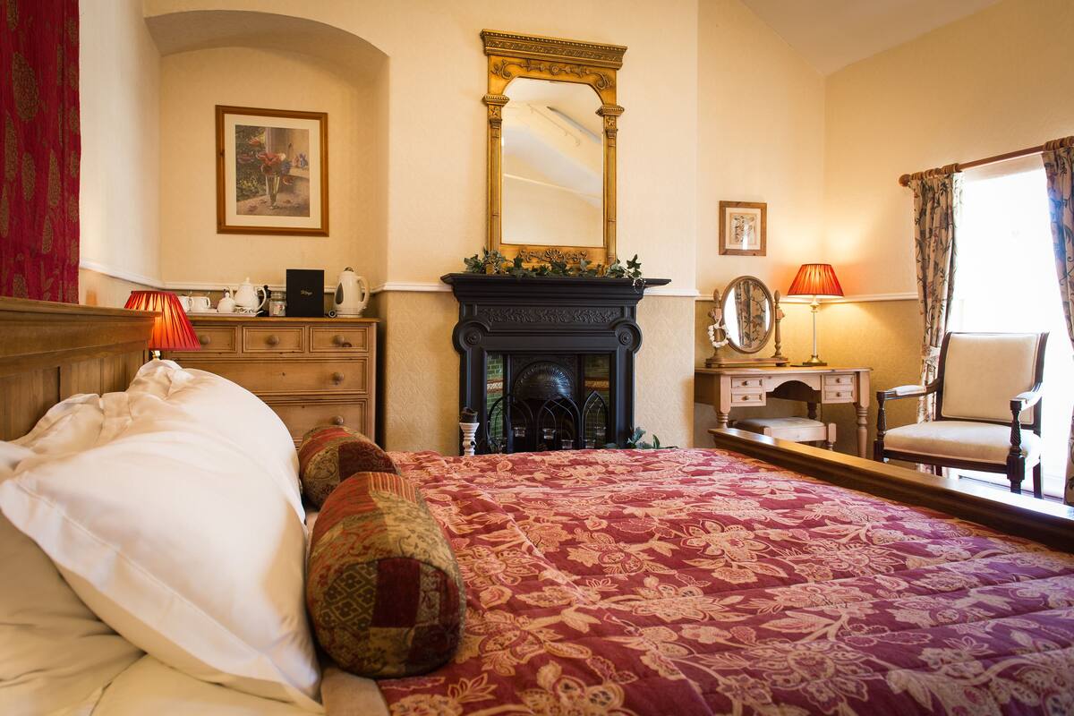A bedroom with a large bed, a fireplace, a mirror, and a desk with a chair.