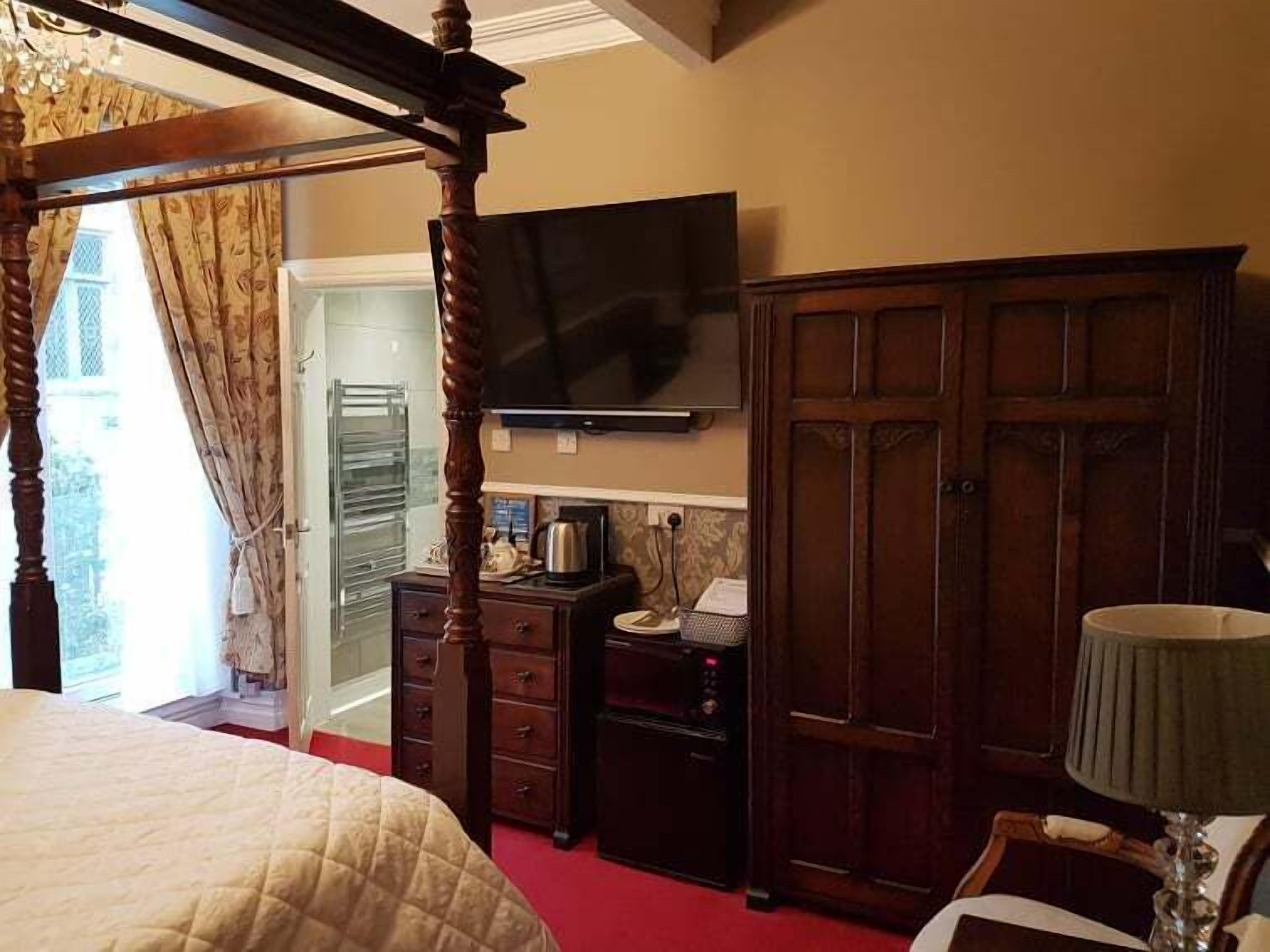 A bedroom with a four-poster bed, a television mounted on the wall, a wardrobe, and a desk with a kettle and papers.