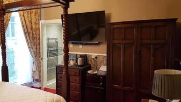 A bedroom with a four-poster bed, a television mounted on the wall, a wardrobe, and a desk with a kettle and papers.