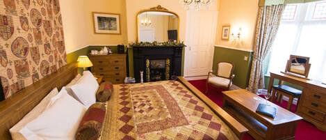 A traditional room with a large bed, a fireplace, a wooden dresser, a chair, and a desk.