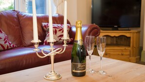 A bottle of Freixenet champagne, two glasses, and a candelabra with three white candles on a wooden table in a living room with a red leather sofa and a television in the background. - Victoria House Room Only Accommodation (Caernarfon)