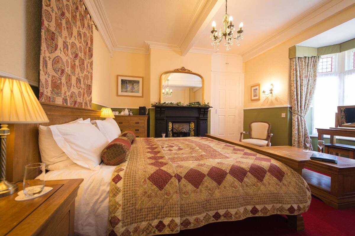 A hotel room with a large bed, a wooden nightstand, a desk, a chair, a fireplace, and a chandelier.