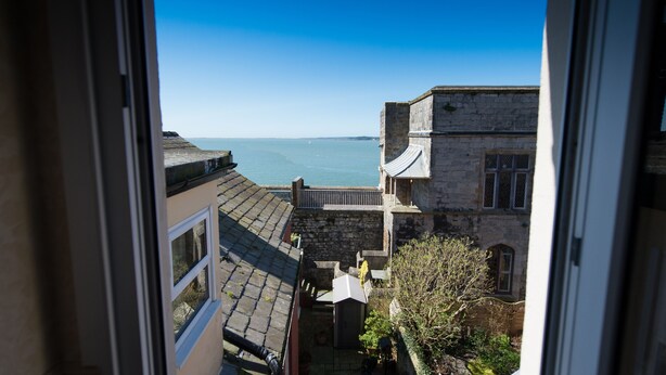 A view from a window overlooking a stone building with a balcony, a clear blue sky, and a calm sea. - Victoria House Room Only Accommodation (Caernarfon)