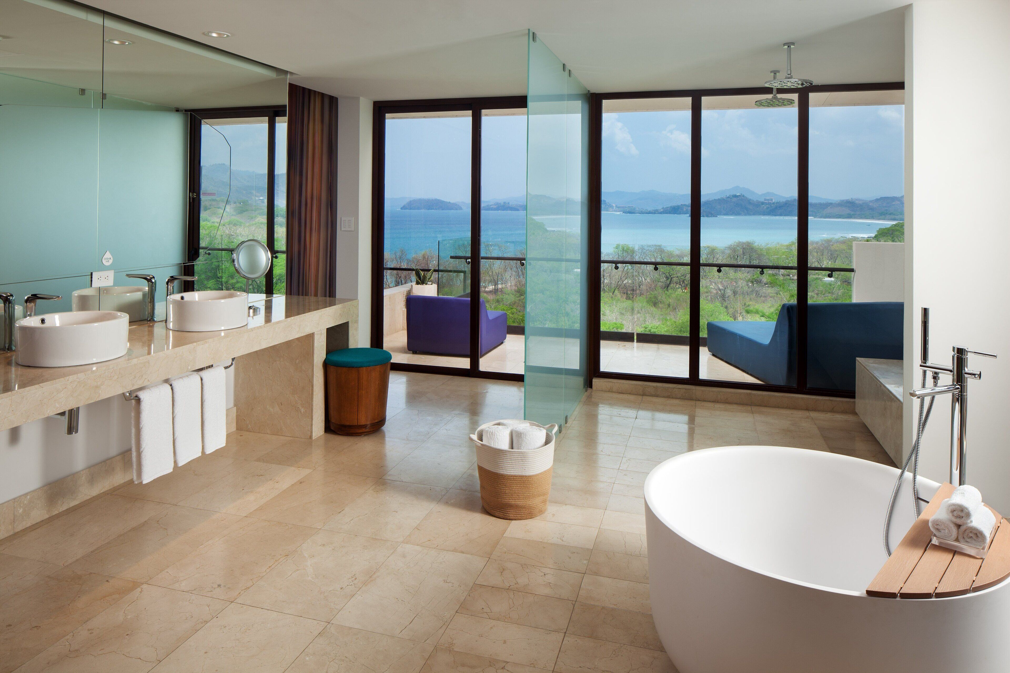 A modern bathroom with a large bathtub, two sinks, and a large window offering a view of the sea.