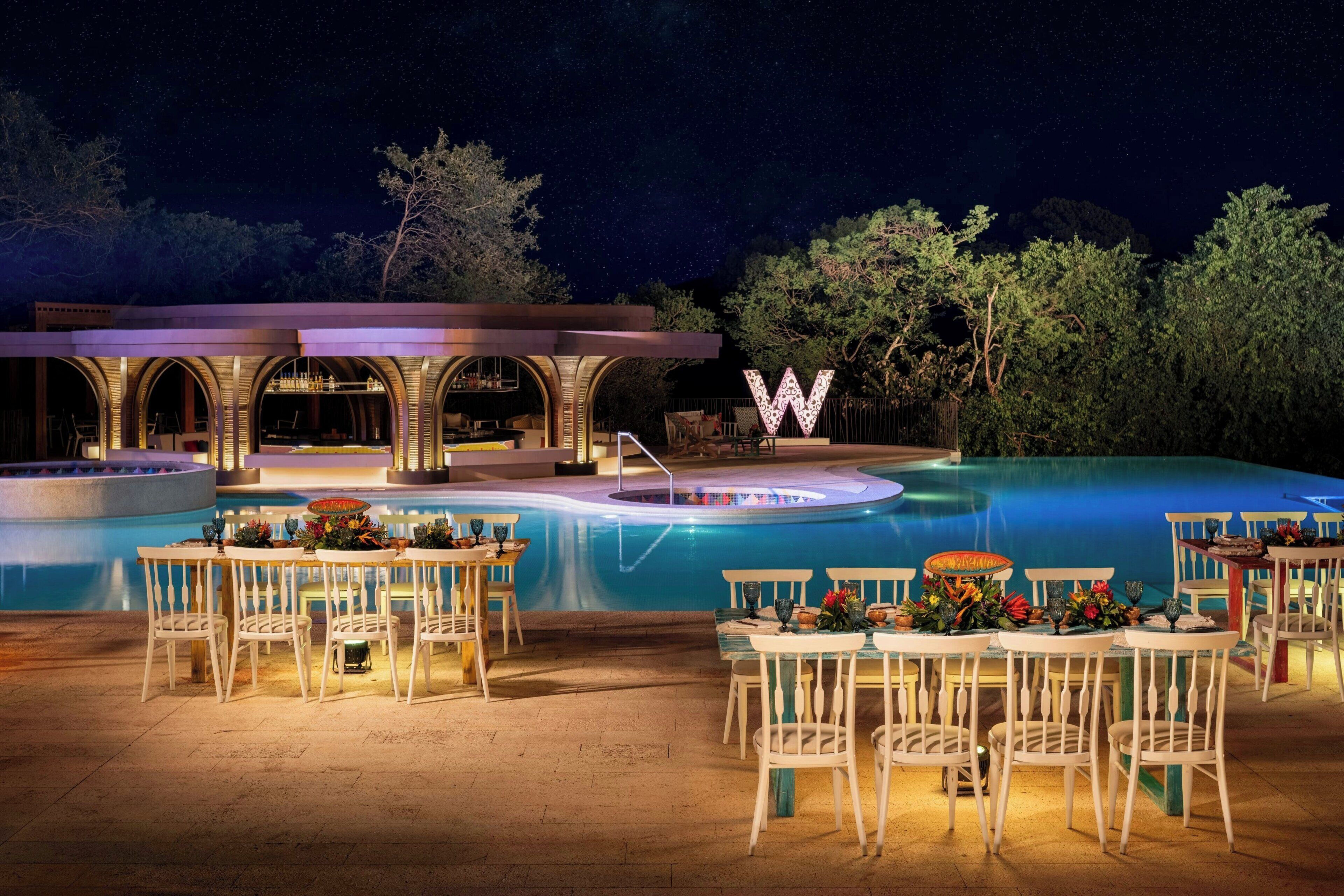 Un área de piscina al aire libre con mesas y sillas, un gran letrero iluminado con la letra 'W', y una zona de estar cubierta.