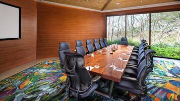 Un salón de conferencias con una larga mesa, varias sillas, una pantalla de proyector y grandes ventanales con vistas a árboles.